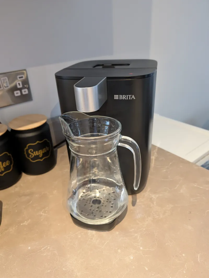 Glass jug placed on the base of a black BRITA dispenser under the spout, with the drip tray removed to make room