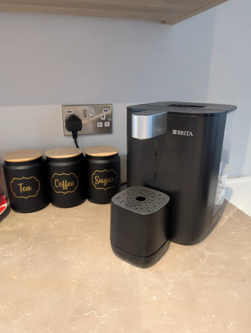 Black BRITA YOURCE water dispenser with silver spout on a stone-effect kitchen worktop, beside tea, coffee and sugar canisters